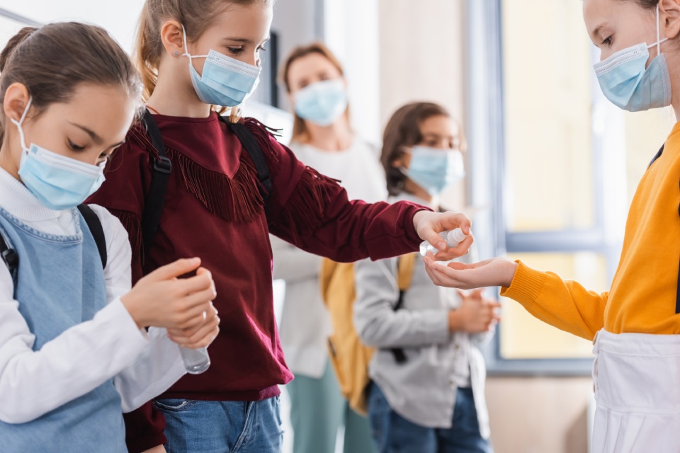 Niños con mascarilla en un colegio se ayudan entre ellos a desinfectarse las manos con esprays